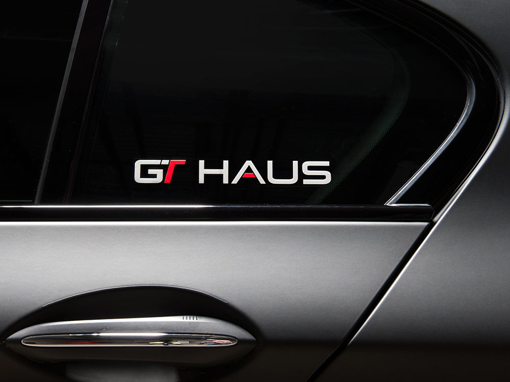GTHAUS decal displayed on a car window, featuring bold lettering in red and silver on a transparent background.