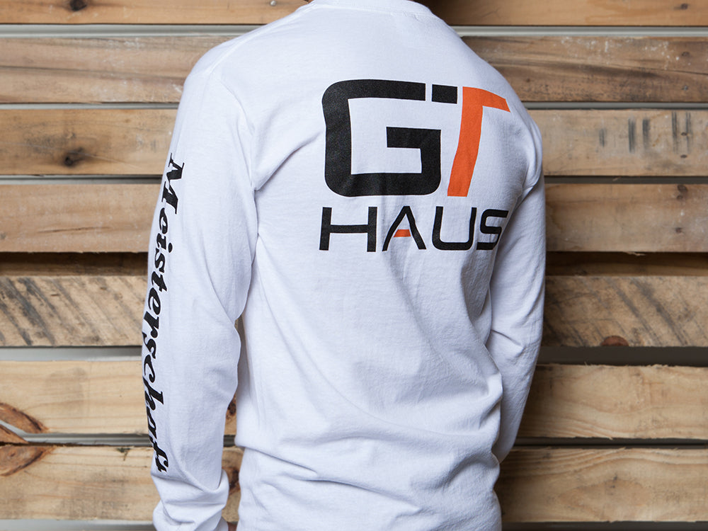 GTHAUS long-sleeve T-shirt featuring a bold logo on the back and text along the sleeve, set against a wooden backdrop.