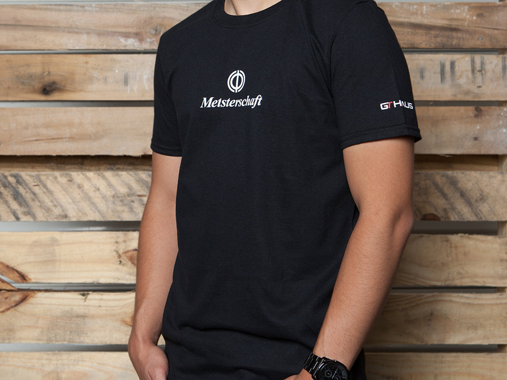 Black MEISTERSCHAFT T-shirt featuring logo, designed for BMW M3 enthusiasts, displayed against a wooden backdrop.