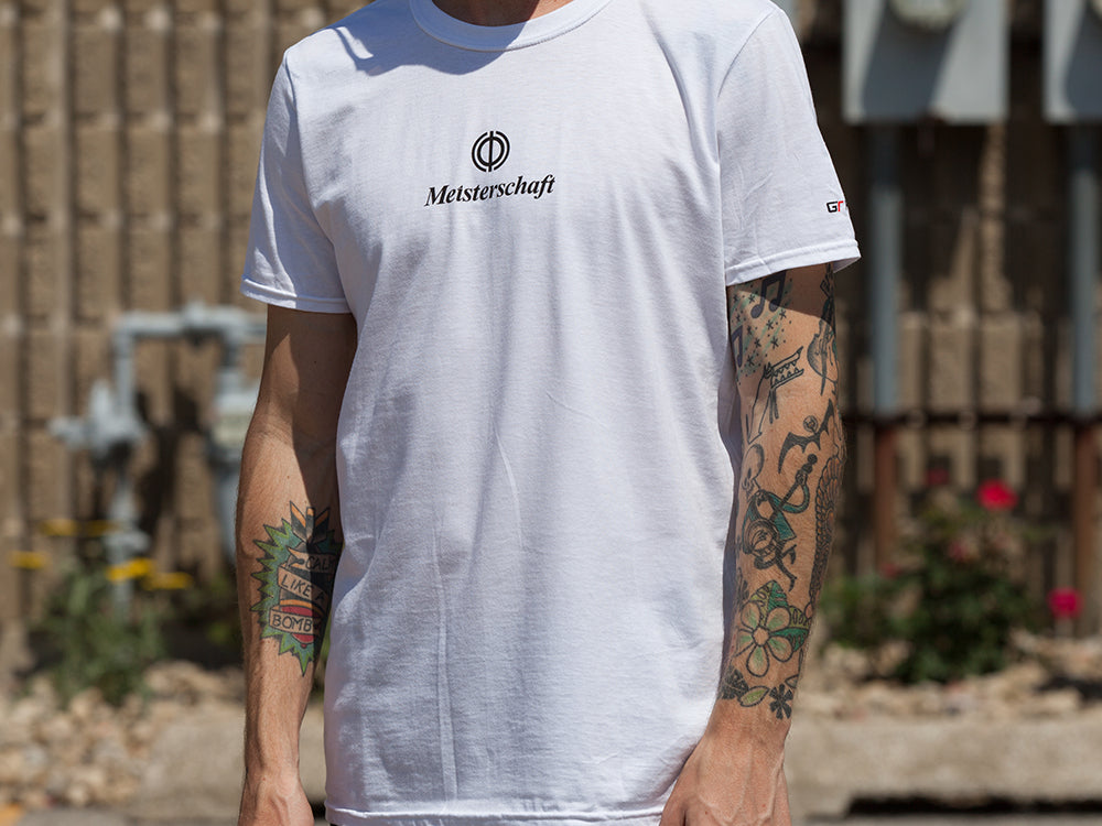 White MEISTERSCHAFT T-shirt featuring a logo, worn by a person with tattoos, set against an outdoor background.