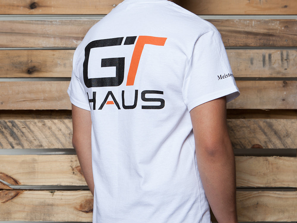 White GTHAUS T-shirt featuring a bold black and orange logo on the back, displayed against a wooden backdrop.