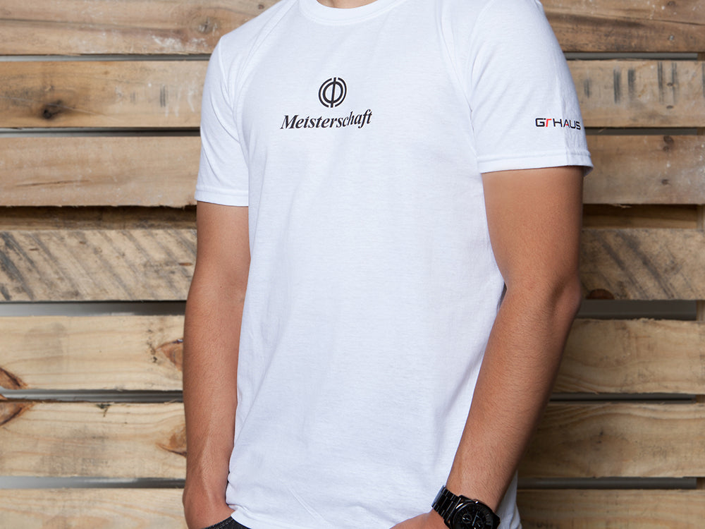 White MEISTERSCHAFT T-shirt featuring a logo, worn by a person against a wooden backdrop, showcasing automotive style.
